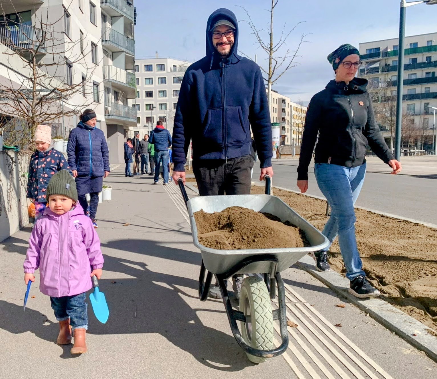 Eine Familie mit Kind zieht mit einer Scheibtruhe aus, um den öffentlichen Raum in der Seestadt zu begrünen